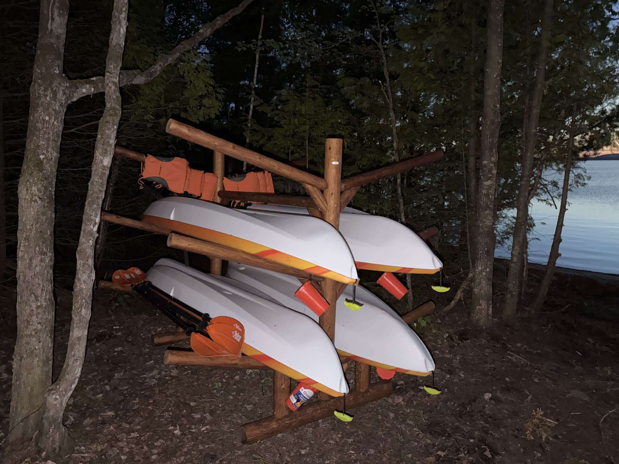 Un support en bois sur lequel sont disposés plusieurs kayaks blancs, rehaussés de touches orange et jaunes, le tout se détachant sur un arrière-plan composé d’arbres et d’une étendue d’eau.