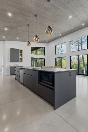 A modern kitchen with a large island, pendant lighting, and large windows offering views of the outdoors.