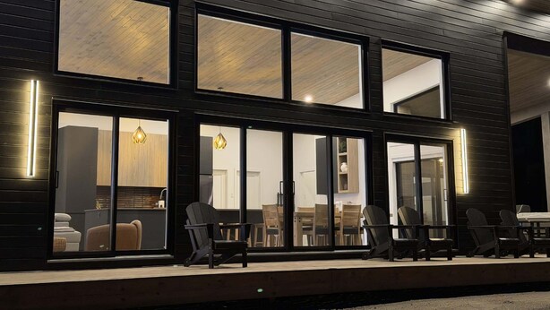 A modern building with large windows, a deck with chairs, and a visible interior with a dining area.