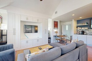 Uma sala de estar moderna com uma grande televisão de tela plana, sofás cinzas, uma mesa de centro de madeira e uma área de jantar com uma mesa e cadeiras.