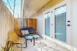 Eine überdachte Terrasse mit Holzlattenzaun, einem Korbstuhl, einem blauen Sofa mit Kissen und einem kleinen Tisch.