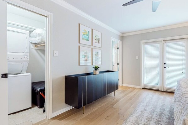 Ein Zimmer mit Einbauwaschmaschine und Trockner, einem schwarzen Schrank mit Pflanze, gerahmten Bildern an der Wand und einem großen Fenster mit Jalousien.
