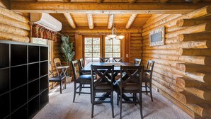 Interior de una cabaña de troncos con una mesa de comedor, sillas, un mueble negro para entretenimiento y un ventanal grande.