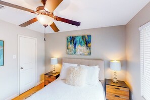 A bedroom with a bed, two bedside tables with lamps, a ceiling fan, a door, and a painting on the wall.