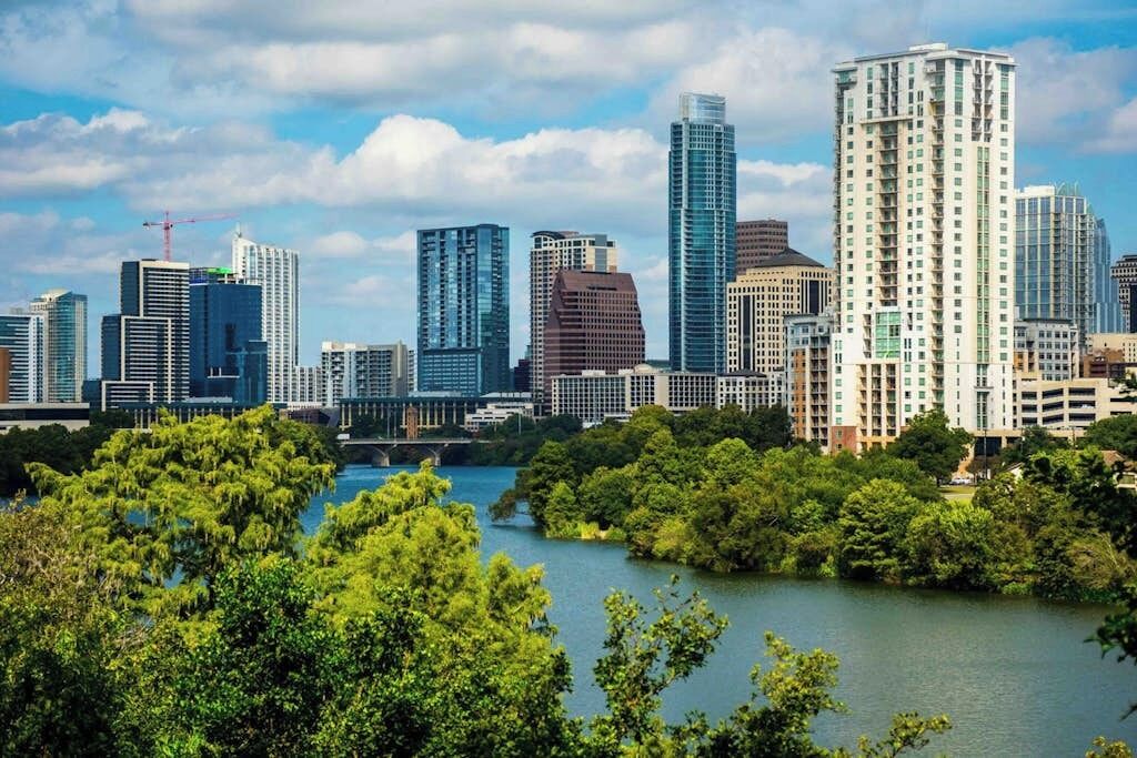 A city skyline with modern high-rise buildings, a river, and lush greenery in the foreground.