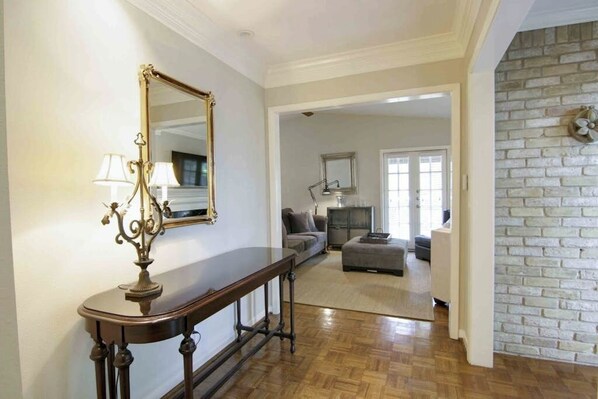 A hallway with a wooden console table, a mirror, and a lamp. There is a sofa and ottoman in the background.
