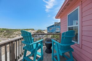 Eine Holzterrasse mit türkisfarbenen Stühlen und einem Tisch, mit Blick auf einen Strand und ein blaues Haus mit einem Fenster.