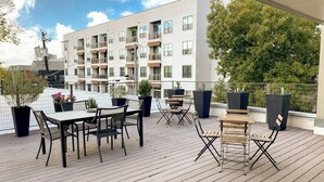 Una terraza en la azotea con una mesa y sillas, plantas en macetas y un edificio moderno al fondo.