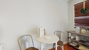 Un coin repas comprenant une table ronde, deux chaises, une petite étagère avec quelques objets et une fenêtre avec des stores et une guirlande décorative.
