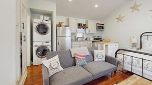 Un espace de vie compact comprenant un coin cuisine, un canapé agrémenté de coussins décoratifs, un lit avec un cadre en métal et un lave-linge.