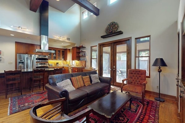 A spacious living room with a sofa, armchair, and coffee table, a kitchen with modern appliances, and large windows allowing natural light. - Charming home with balcony, firepit, & 2 living rooms - near dining & music (Austin)