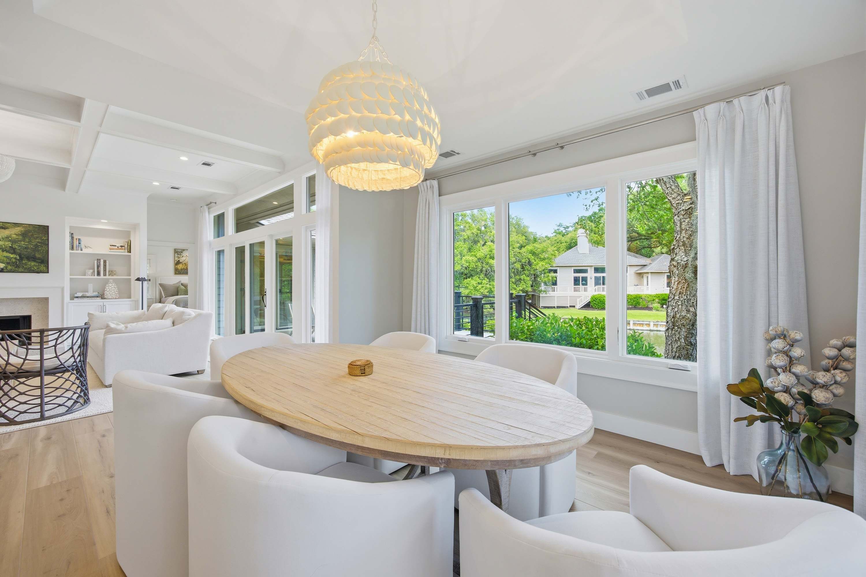 A modern interior with a round wooden table surrounded by white chairs, large windows with curtains, and a hanging light fixture.