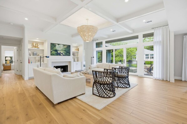 Ein geräumiges Wohnzimmer mit Kamin, weißen Sofas, einer Chandelier und großen Fenstern.