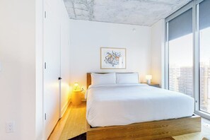 Un dormitorio moderno con una cama plataforma de madera, vistas a la ciudad, una obra de arte enmarcada en la pared y una lámpara de noche.
