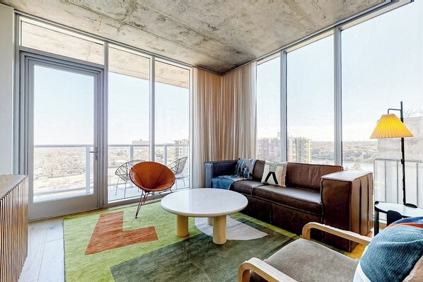 Una sala de estar moderna con un sofá, una mesa de centro redonda y un sillón. Una ventana grande ofrece vistas de la ciudad.