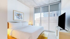 Une chambre d’hôtel moderne dotée d’un grand lit, d’un téléviseur à écran plat, d’une fenêtre donnant sur les bâtiments et d’une œuvre d’art abstraite encadrée accrochée au mur.