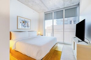 A modern hotel room with a large bed, a flat-screen TV, a window with a view of buildings, and a framed abstract artwork on the wall. - 13th floor corner condo, lake-view balcony, rooftop pool & gym, dog-friendly (Austin)