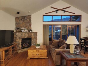 Ein Wohnzimmer mit Kamin aus Stein, Holzmöbeln, einem Fernseher und großen Fenstern mit Blick ins Freie.