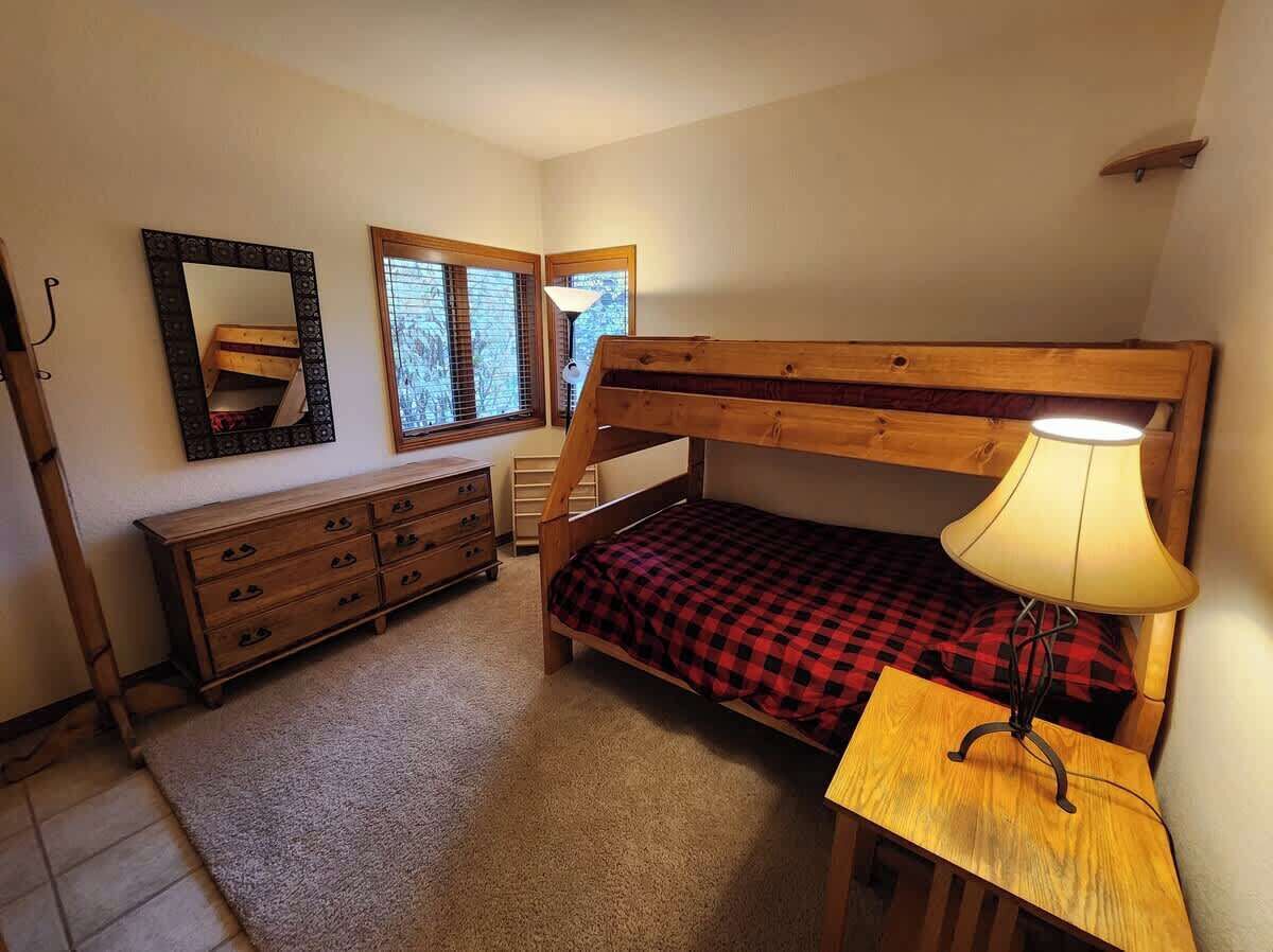 A bunk bed with a lamp, a dresser, a mirror, and a window with blinds.