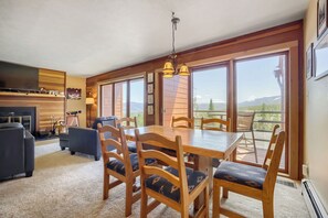 Ein geräumiger Wohnbereich mit einem großen Esstisch aus Holz, gemütlicher Sitzgelegenheit, Kamin und einem Balkon mit Bergblick.