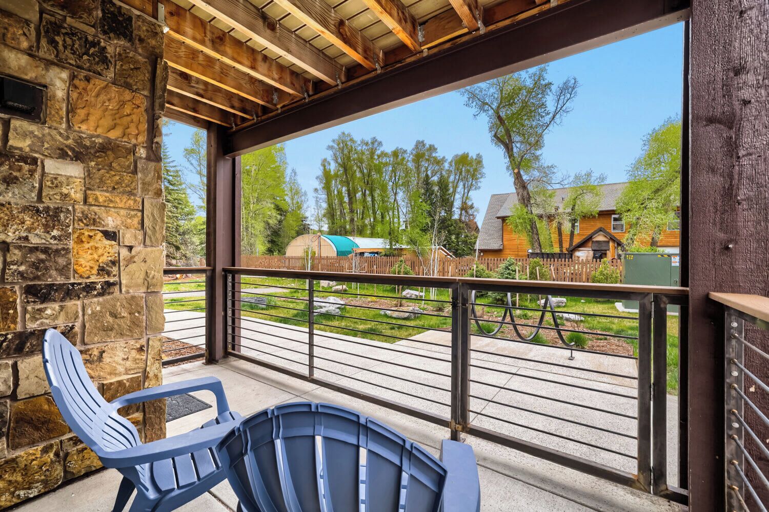 Un espacio al aire libre techado, con pared de piedra, techo de madera y vistas a una zona residencial con casas y árboles.