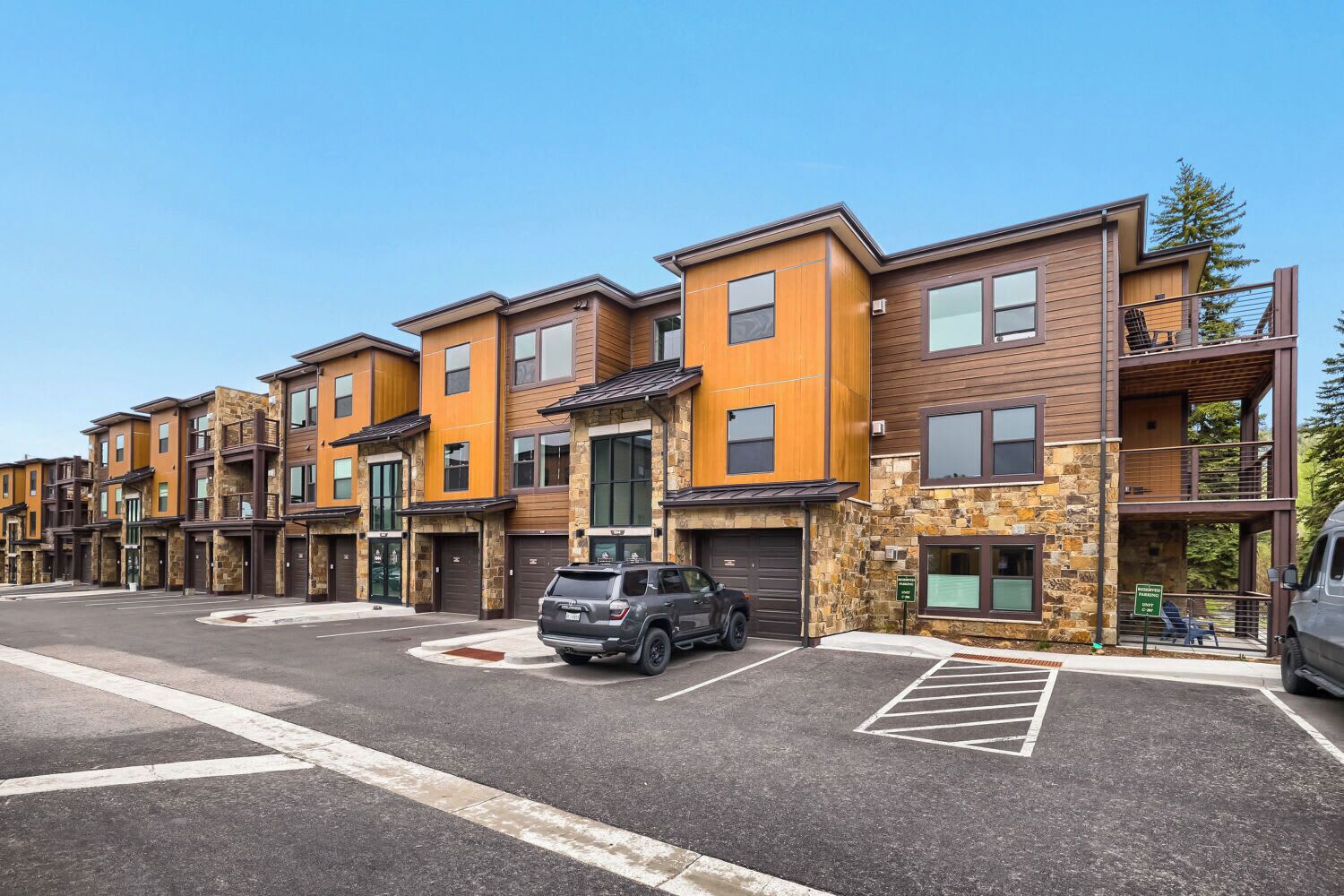 A row of modern multi-story apartment buildings with stone and wood exteriors, a parking area, and a clear sky.