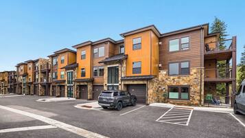 A row of modern multi-story apartment buildings with stone and wood exteriors, a parking area, and a clear sky.