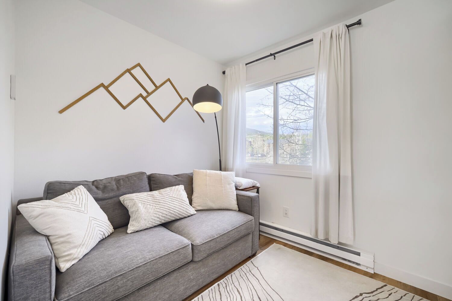 A modern living room with a grey sofa, white cushions, a geometric wall art piece, a floor lamp, and a large window with curtains.