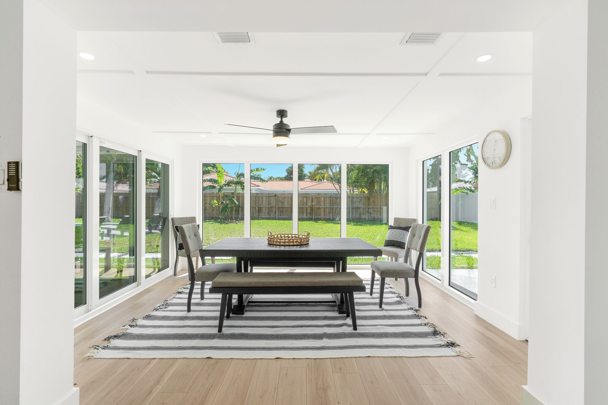 A dining area with a table, chairs, a bench, a ceiling fan, and large windows with a view of a backyard.