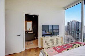 A bedroom with a large window, a bed, a TV, and a dresser. - Luxe Rainey St corner condo with lakeview balcony, rooftop pool & gym (Austin)