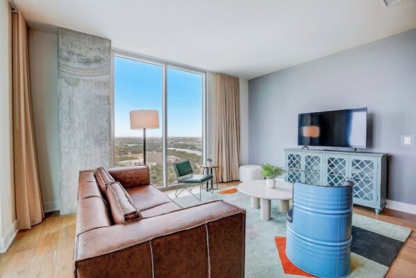 A modern living room with a sofa, a television, a cabinet, and a large window. - Luxe Rainey St corner condo with lakeview balcony, rooftop pool & gym (Austin)