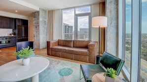 Ein modernes Wohnzimmer mit einer Couch, einem Esstisch und einem Fernseher. Eine Küchenzeile mit dunklen Schränken und einem Kühlschrank ist ebenfalls vorhanden. Große Fenster bieten Blick auf die Stadtlandschaft.