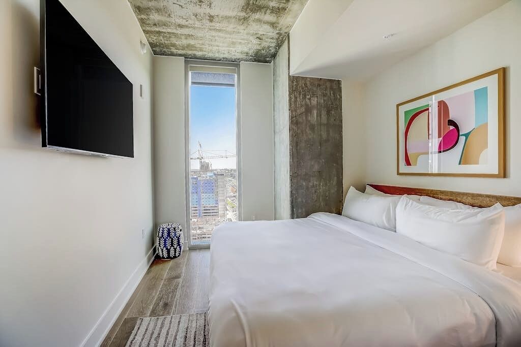 Una habitación de hotel con una cama grande, un televisor de pantalla plana en la pared, un ventanal con vistas a edificios y una obra de arte abstracto en la pared.
