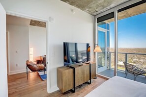 Moderni hotellihuone, jossa on suuri televisio, puinen konsolipöytä, parveke, jolta on näkymä, ja viihtyisä oleskelualue.
