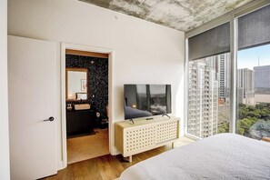 Ein Schlafzimmer mit einem Bett, einem Fernseher auf einem Schrank, einem Badezimmer mit Spiegel und Blick auf Gebäude durch ein großes Fenster.