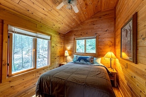Une chambre dans une cabane en bois, avec un lit, deux fenêtres, un ventilateur de plafond et une table de chevet avec une lampe.