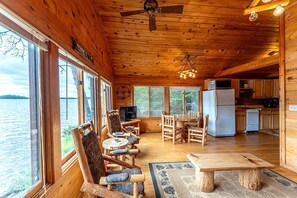 A cabin with wooden walls, a dining area with a table and chairs, a kitchen with a refrigerator and stove, and large windows overlooking a body of water. - Secluded Pet-Friendly Cabin Rental on Big Lake for Dreamy Glamping in Ely, MN (Ely)