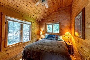 A wooden cabin bedroom with a bed, two windows, a ceiling fan, and a nightstand with a lamp. - Secluded Pet-Friendly Cabin Rental on Big Lake for Dreamy Glamping in Ely, MN (Ely)