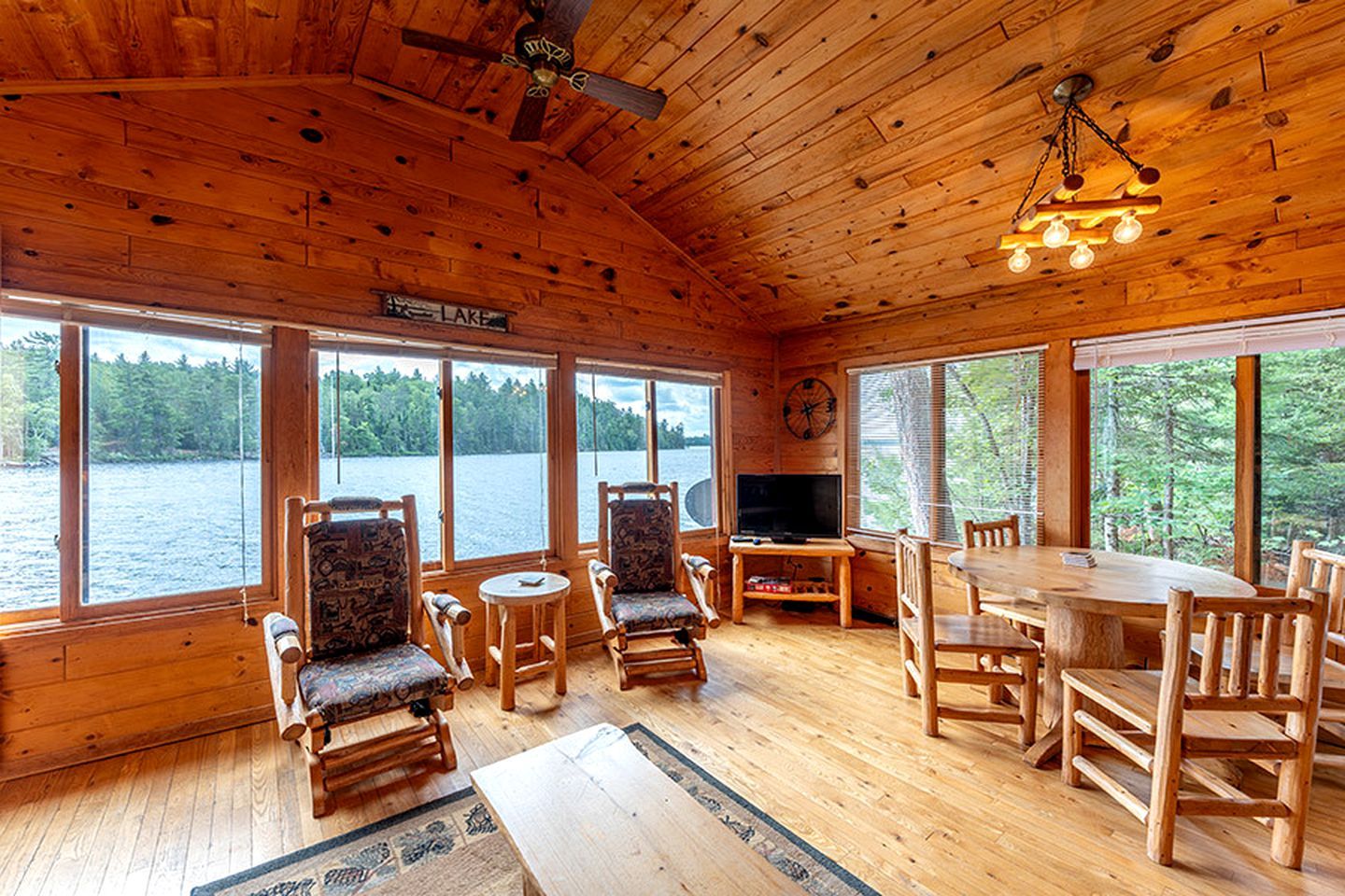 Eine Hütte mit einem großen Holztisch, mehreren Stühlen, einem Fernseher und einer Uhr an der Wand.