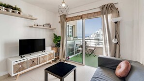 En moderne stue med et fladskærms-tv, en sofa med pude, et sofabord, en balkon med bord og stole, og udsigt over bybilledet.