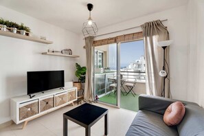 Ein modernes Wohnzimmer mit Flachbildfernseher, einer Couch mit Kissen, einem Couchtisch, einem Balkon mit Tisch und Stühlen und Blick auf die Stadtlandschaft.