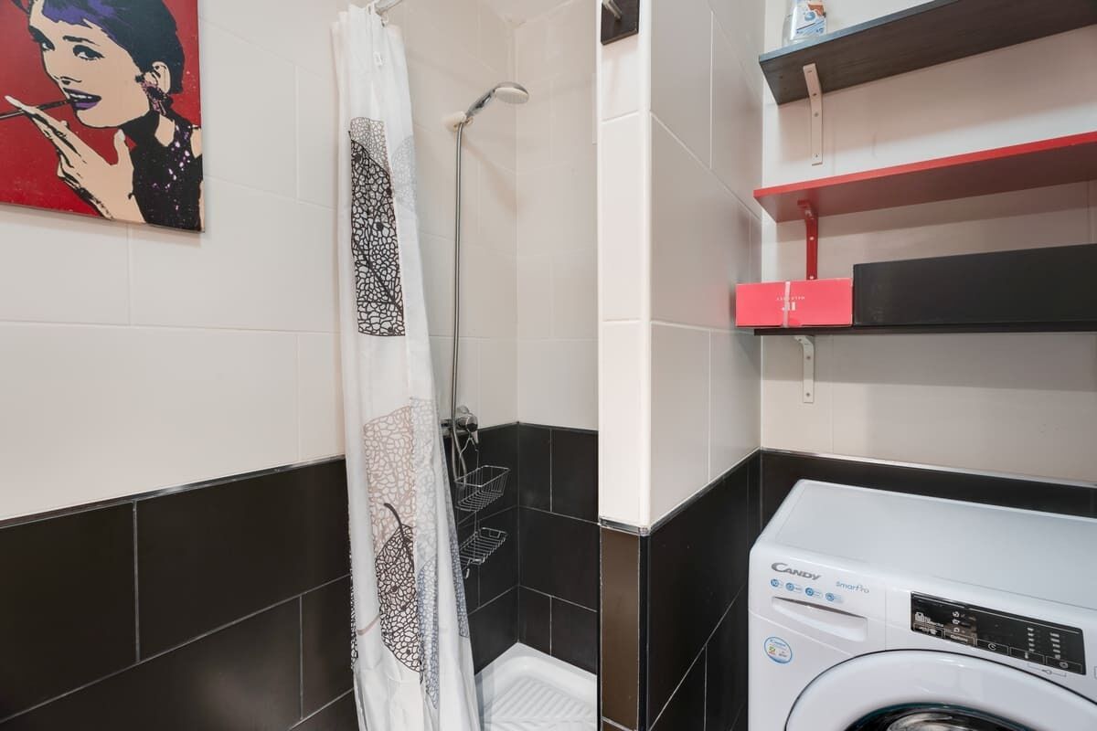 A bathroom with a shower, a toilet, a washing machine, and a shelf with a red box.