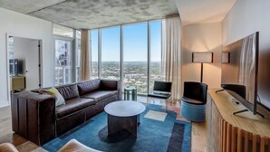 A modern living room with a large window, a leather sofa, a round coffee table, and a TV. - Historic Rainey Street sky-high condo with amazing views & rooftop pool (Austin)