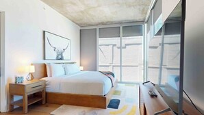 Quarto com cama, mesa de cabeceira, abajur e uma janela grande.