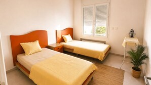 Een slaapkamer met twee bedden, een nachtkastje, een radiator, een raam met zonwering, een klein tafeltje met een lamp en een bloempot met plant.