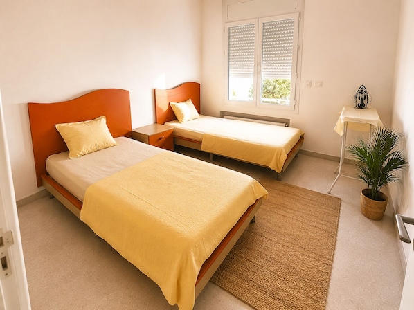 A bedroom with two beds, a bedside table, a radiator, a window with blinds, a small table with a lamp, and a potted plant. - CosyStay Apartment (Tunis)