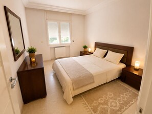 Une chambre à coucher comprenant un lit, des tables de chevet, une commode, un miroir, une fenêtre avec des stores et un radiateur.