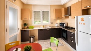 Een moderne keuken met een magnetron, fornuis en koelkast. Er is een raam met zonwering, een prullenbak en een stoel.
