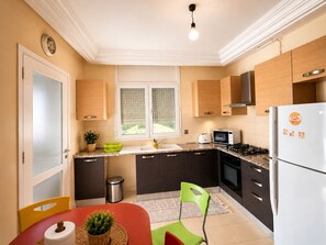 A modern kitchen with a microwave, stove, and refrigerator. There is a window with blinds, a trash can, and a chair. - CosyStay Apartment (Tunis)