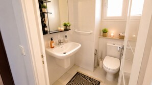 Een badkamer met een wit wastafel, toilet en douche. Boven het wastafel hangt een spiegel, er is een plank met toiletartikelen en een klein raam.